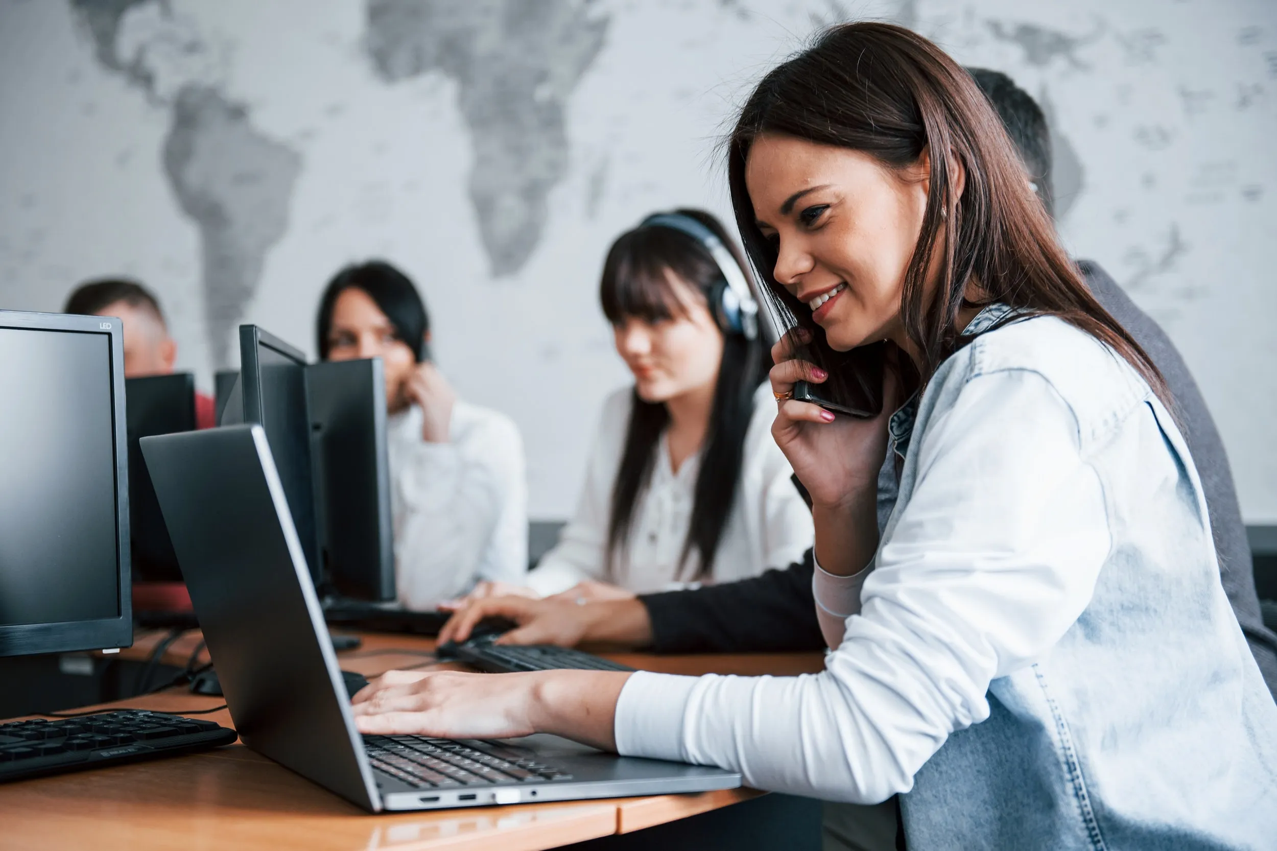 Une femme parle au téléphone tout en utilisant un ordinateur portable dans une salle avec d’autres personnes au travail.